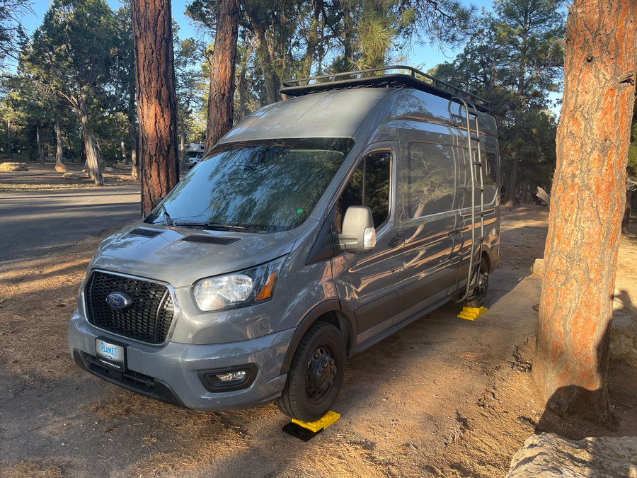 Grey Ford Transit RV rental, leveled on yellow blocks at a sunny forest campsite. Trees surround the vehicle.