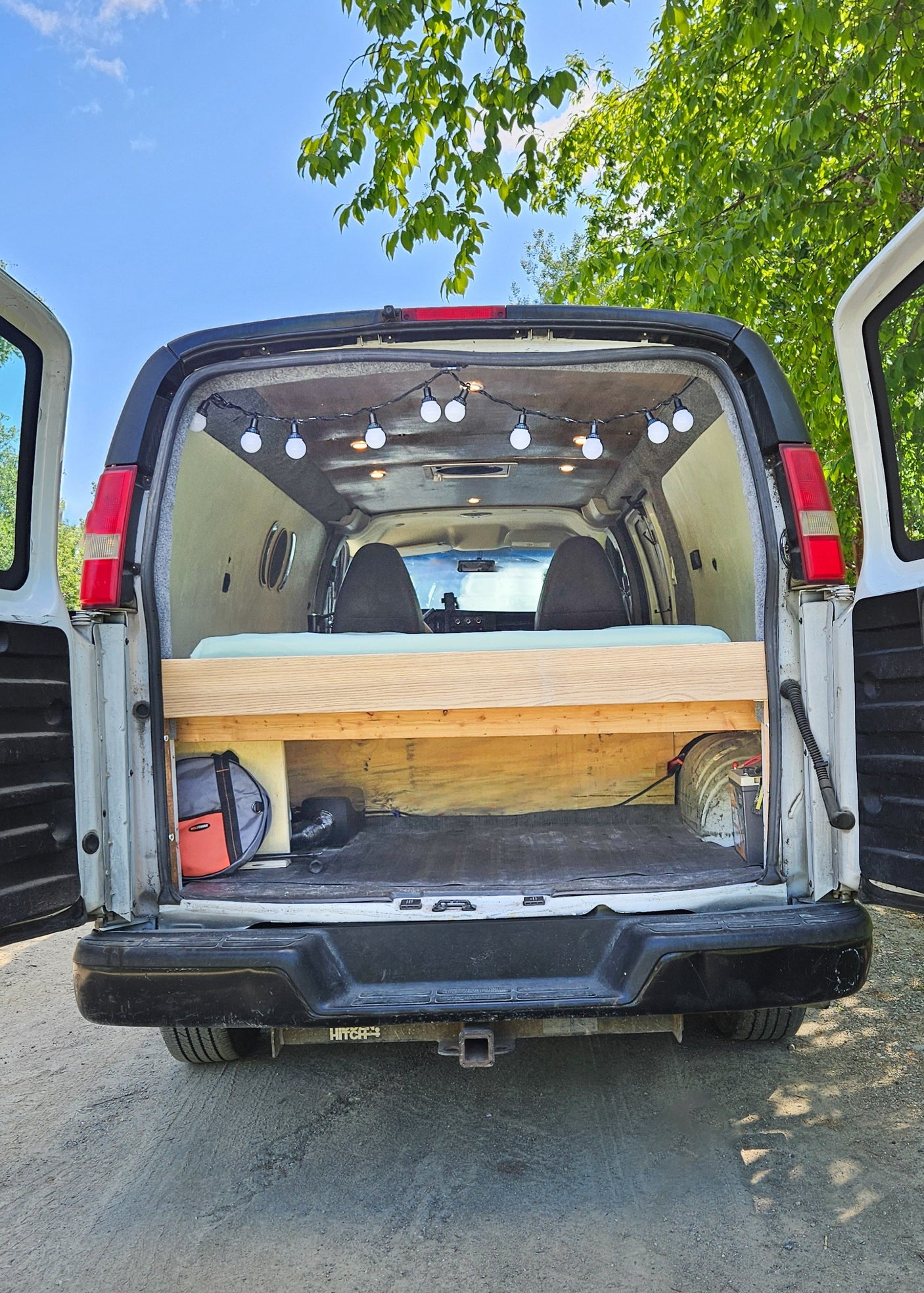 Explore with this custom camper van conversion, ready for adventure! Rear doors open, showing the cozy bed, string lights, and storage in this RV rental.