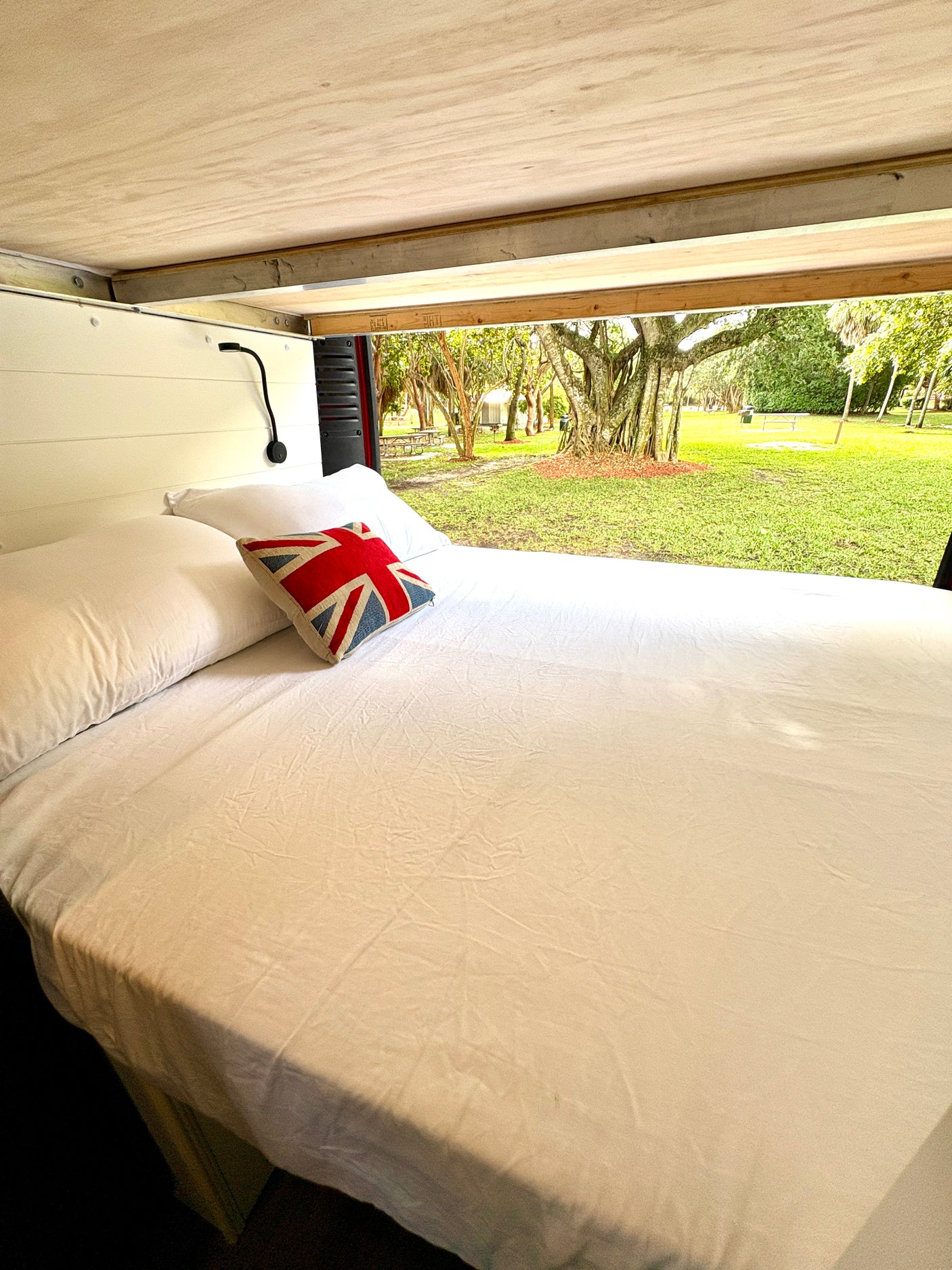 Cozy bed inside an RV rental with white sheets, a Union Jack pillow, and a beautiful outdoor view for your next adventure.