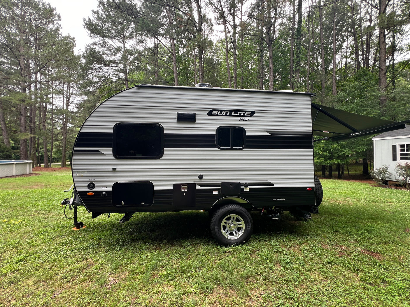 A grey and black striped Sun Lite Sport RV rental, a travel trailer with an extended awning, parked on green grass near trees.