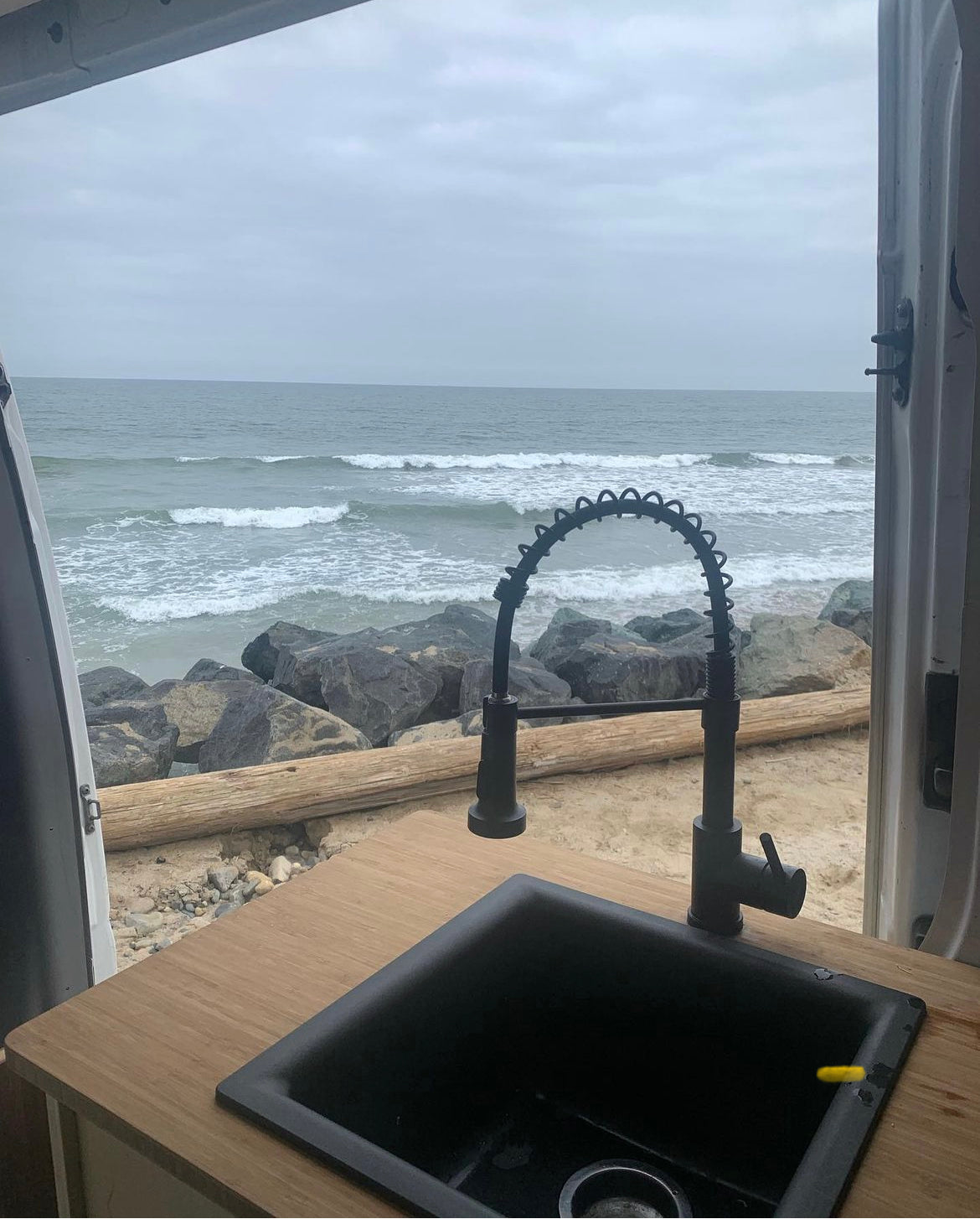 RV rental: Ocean view from a van kitchen with a black sink & faucet, overlooking waves and beach.
