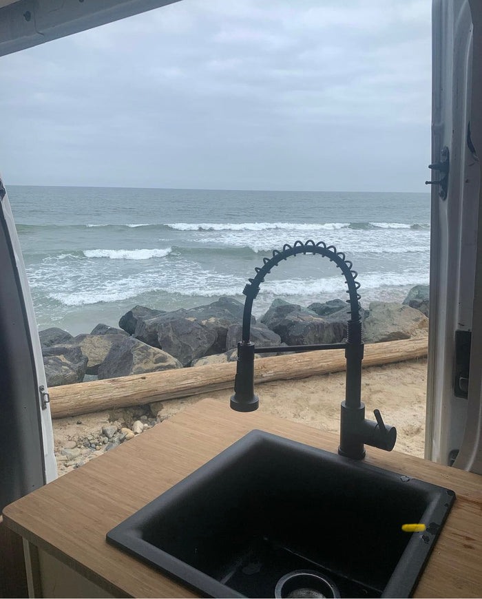 RV rental: Ocean view from a van kitchen with a black sink & faucet, overlooking waves and beach.