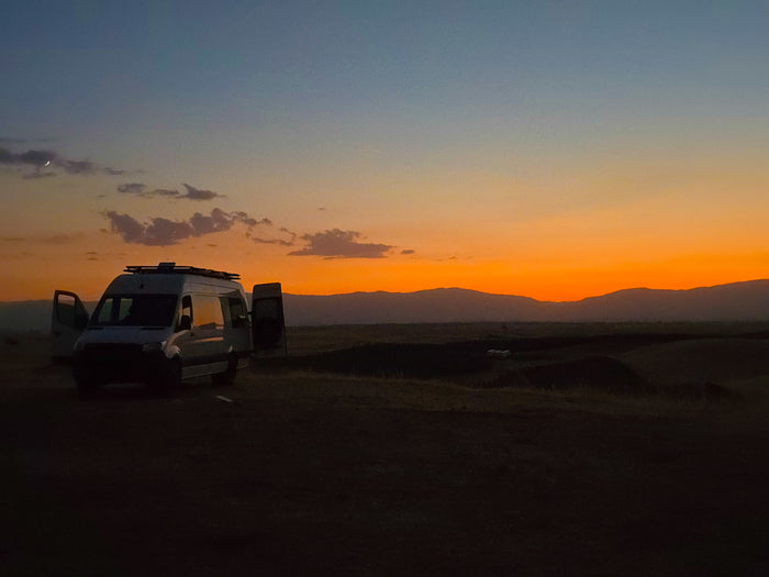 Serene sunset with an RV rental van in a field, mountains, and a crescent moon in the sky.