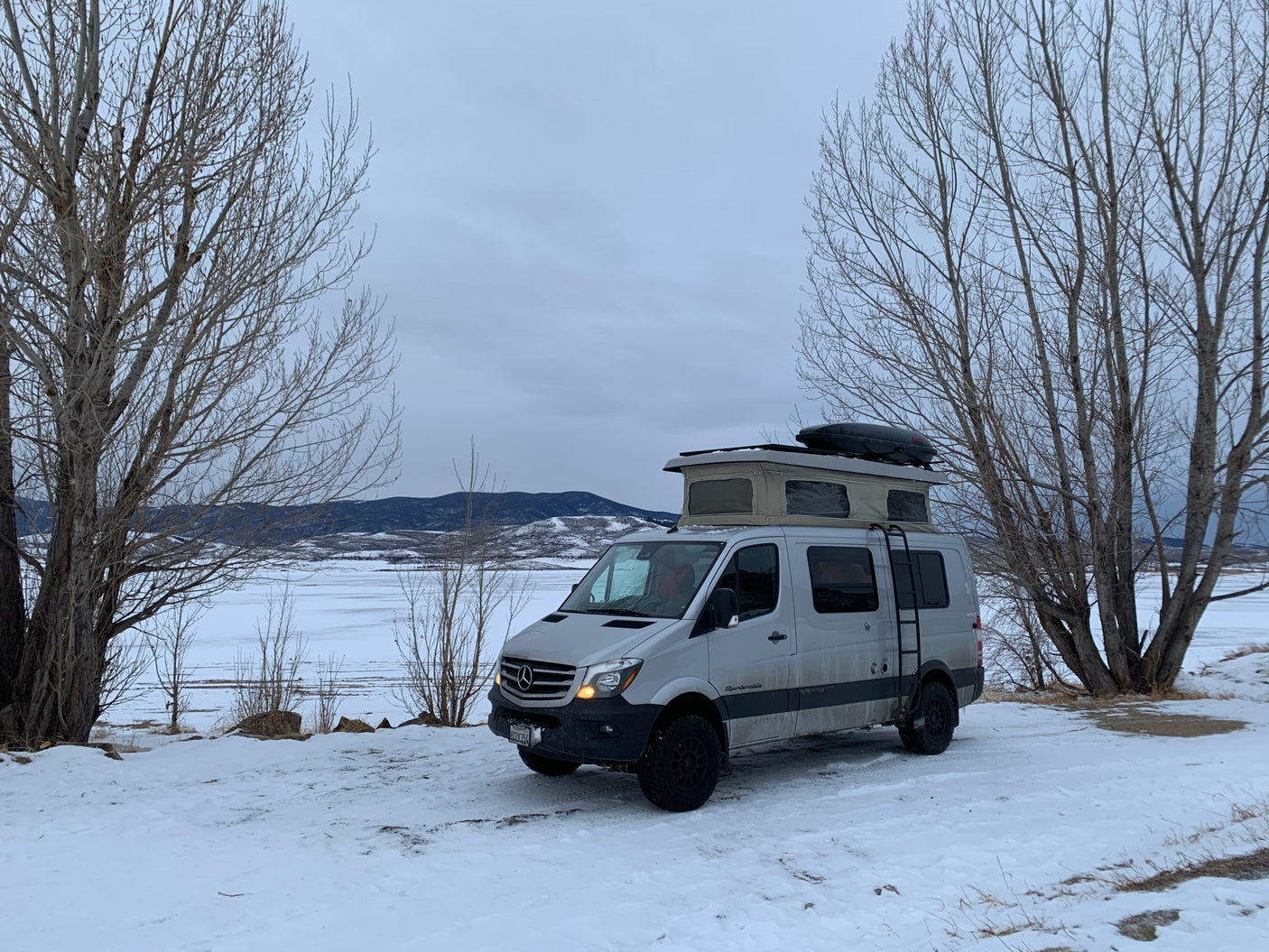 Experience winter adventures with an RV rental! A grey camper van with a pop-top tent is parked on snow beside a frozen lake & mountains.