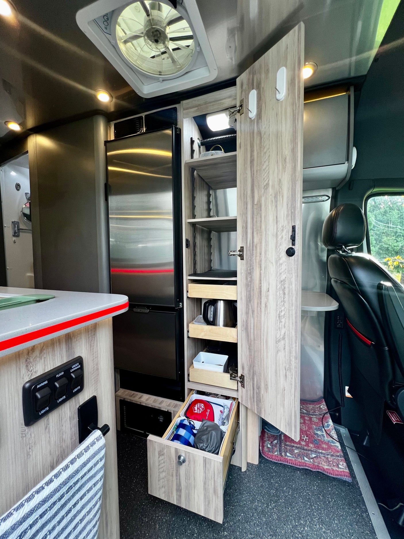 This modern RV rental interior features a stainless steel fridge, an open pantry with pull-out drawers, controls, and a driver's seat.