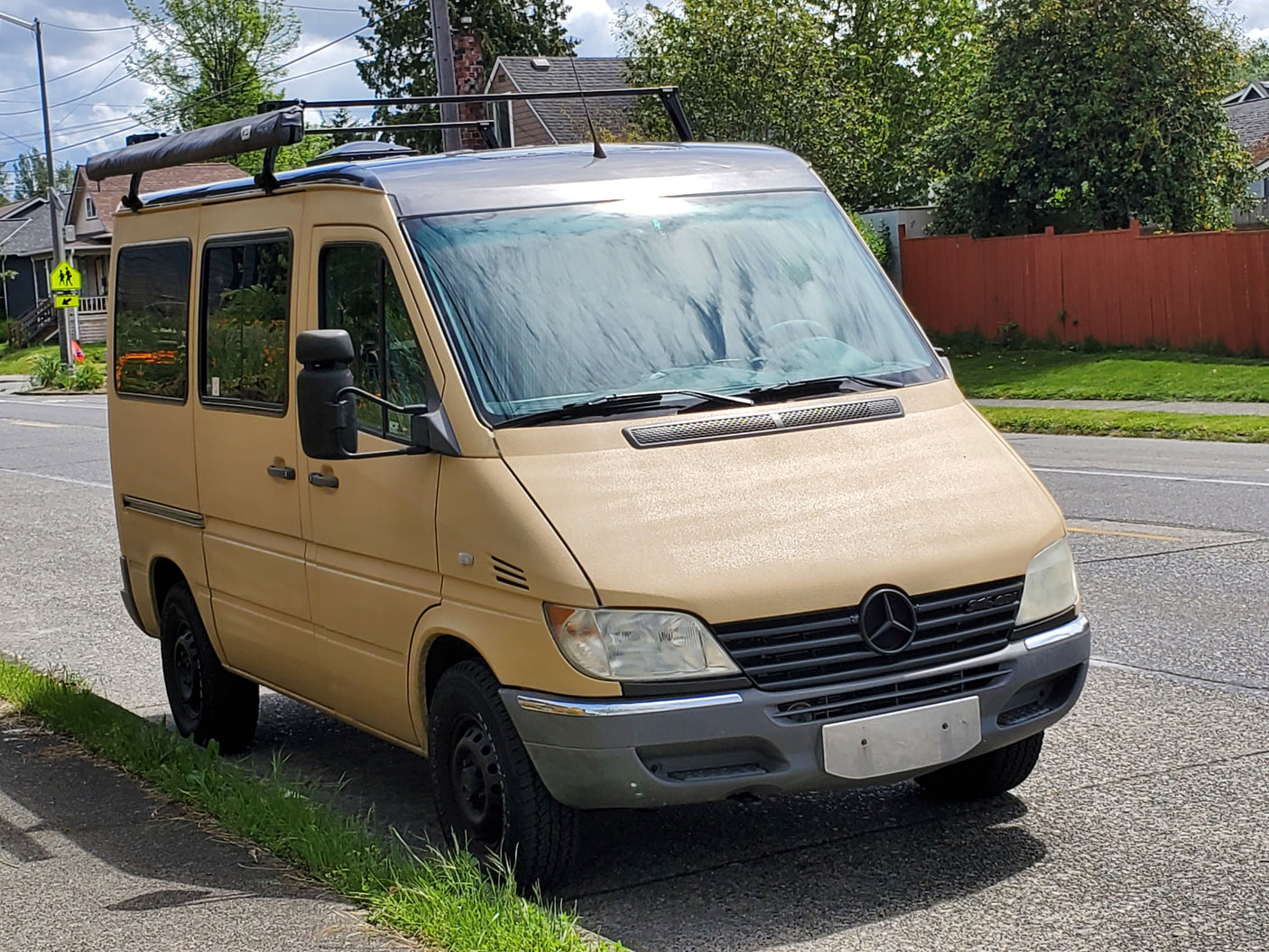 Tan Mercedes-Benz Sprinter van, equipped as an RV rental, parked roadside. Features a roof rack and side awning.