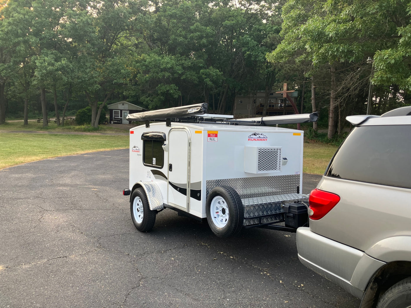Small white Runaway camping trailer, like an **RV rental**, hitched to a silver SUV on pavement in a wooded area. Ready for adventures.