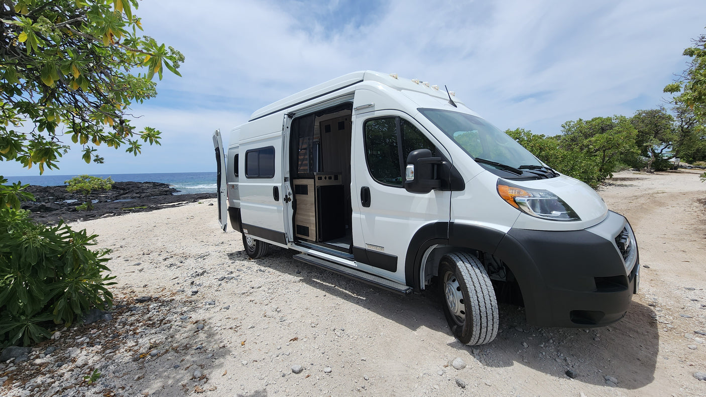 White RV rental camper van on a sandy, rocky beach. Ocean and volcanic shoreline in background. Perfect for nature adventures.