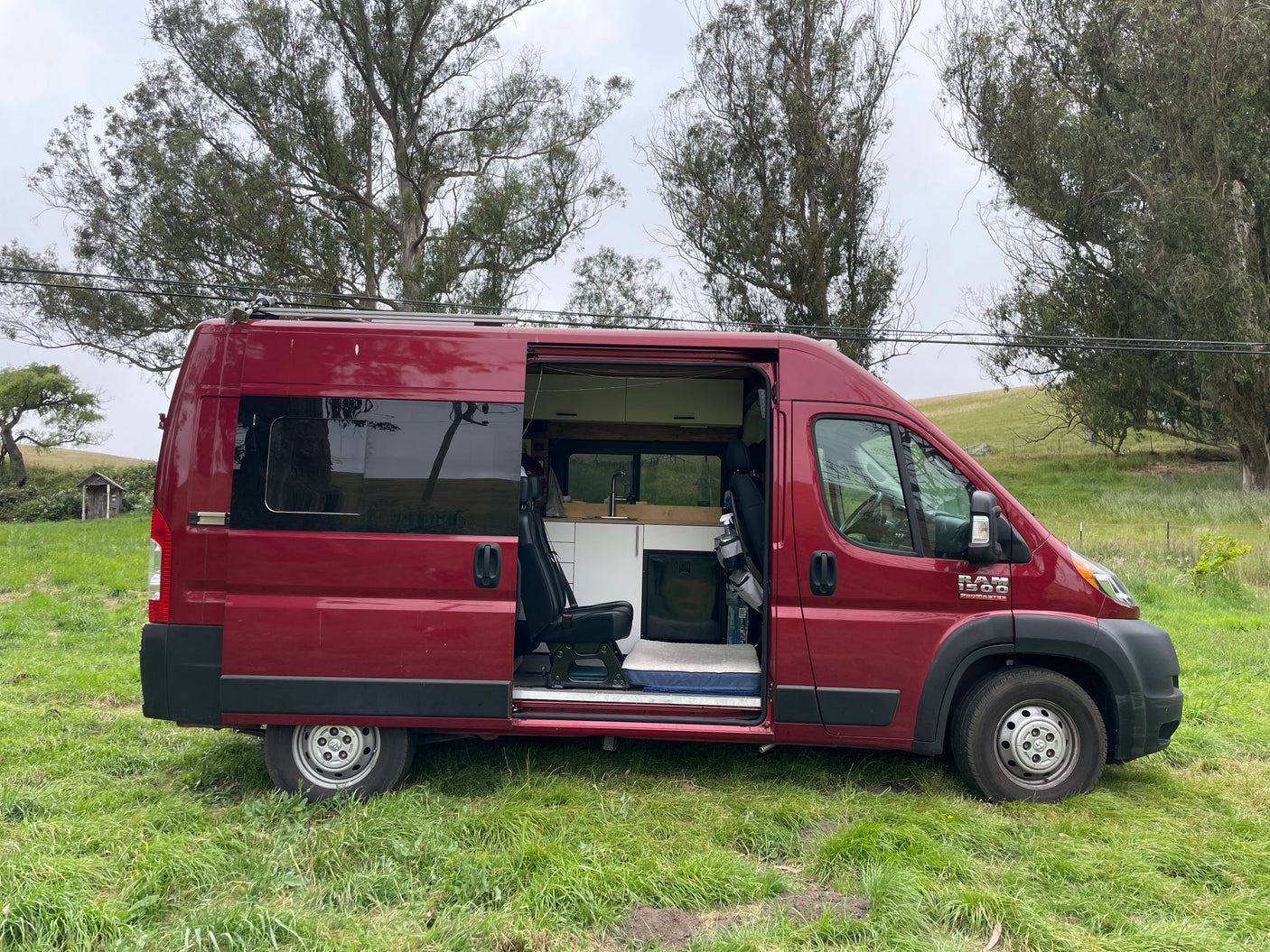 Red Ram ProMaster camper van with open side door showing custom interior for an RV rental adventure in a grassy field.