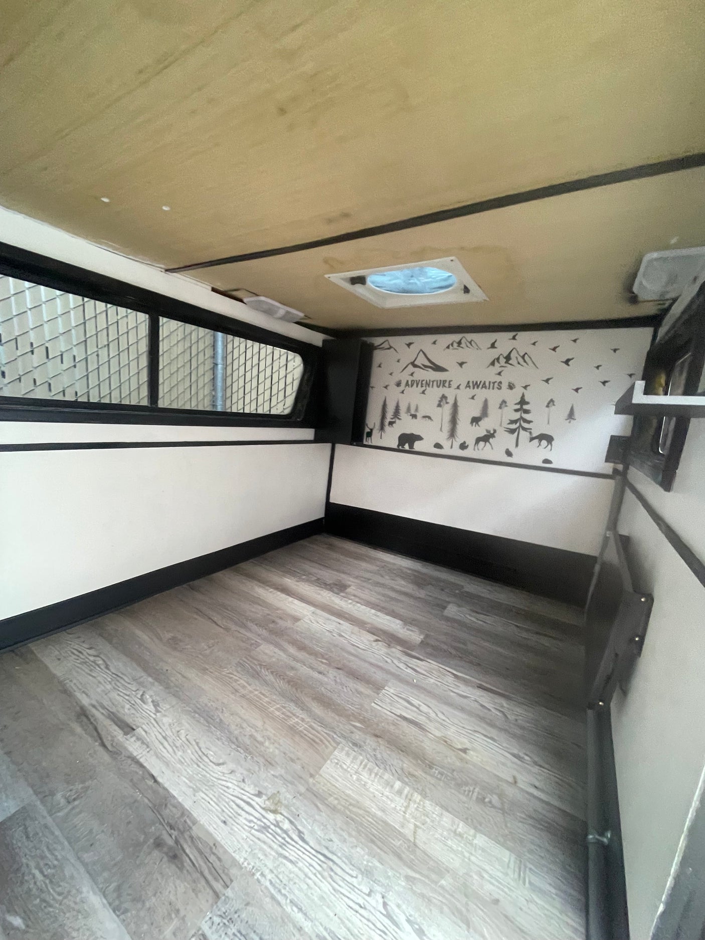 Spacious rv rental interior: wood-look floor, white walls with black trim, and an "ADVENTURE AWAITS" mural.