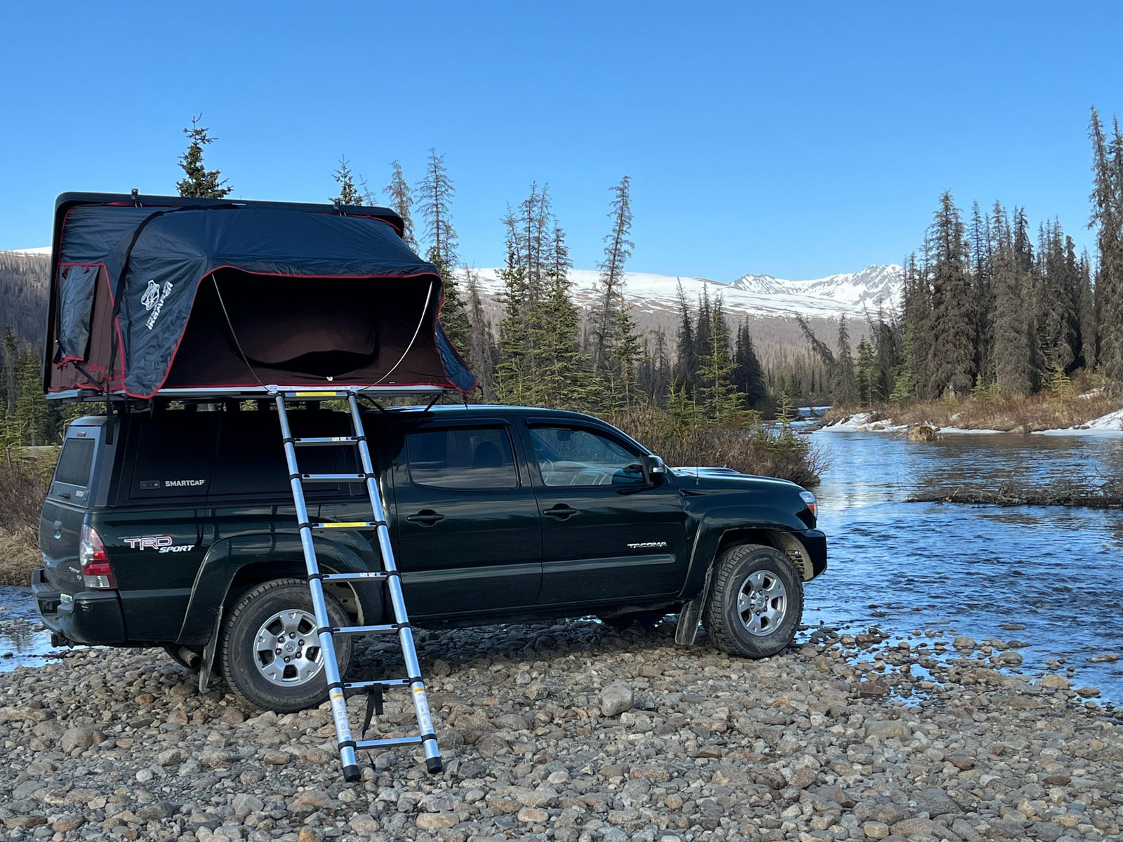 A dark green truck with an open rooftop tent by a river and snow-capped mountains. Ideal for an RV rental outdoor adventure.