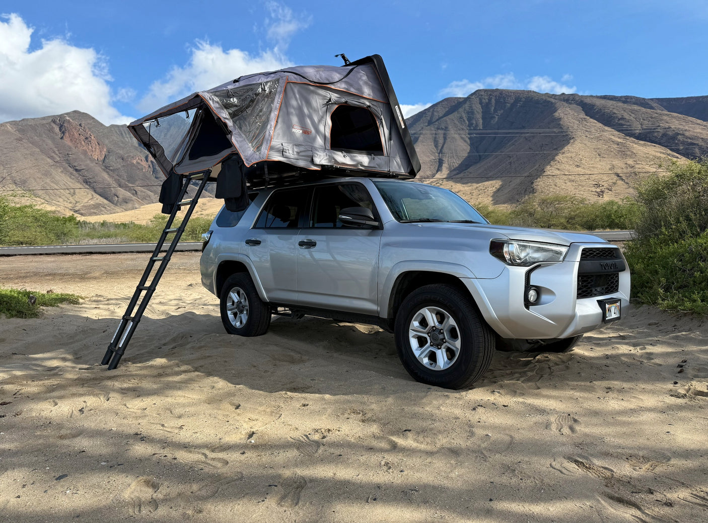 Silver Toyota 4Runner with a deployed Roofnest rooftop tent on sand, mountains behind. Explore with this RV rental.