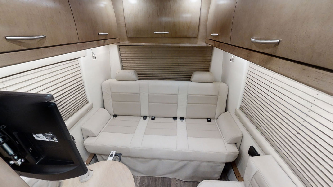 Inside an RV rental: a cream sofa converts to a bed, flanked by blinds & overhead cabinets, with a TV in view.