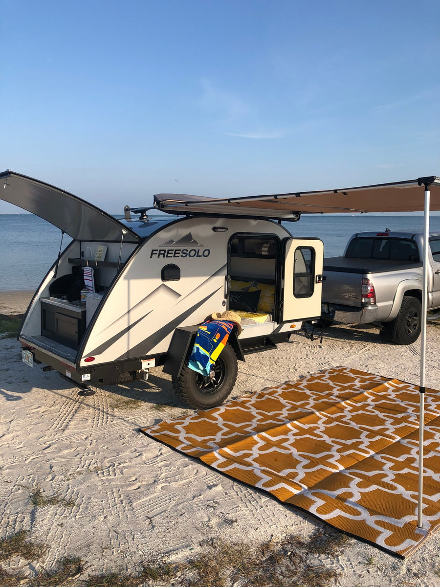 A Freesolo teardrop RV rental is perfectly set up for beach camping. A truck is attached, with an awning and outdoor rug by the ocean.