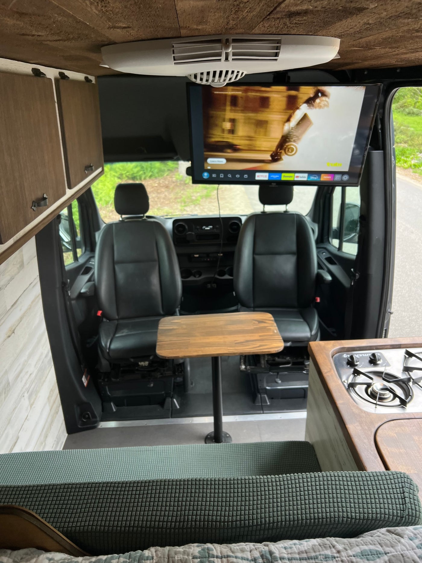 View inside an RV rental showing the front seats, a central table, an overhead TV, a kitchen counter with stove, and wooden cabinets. — photo 6