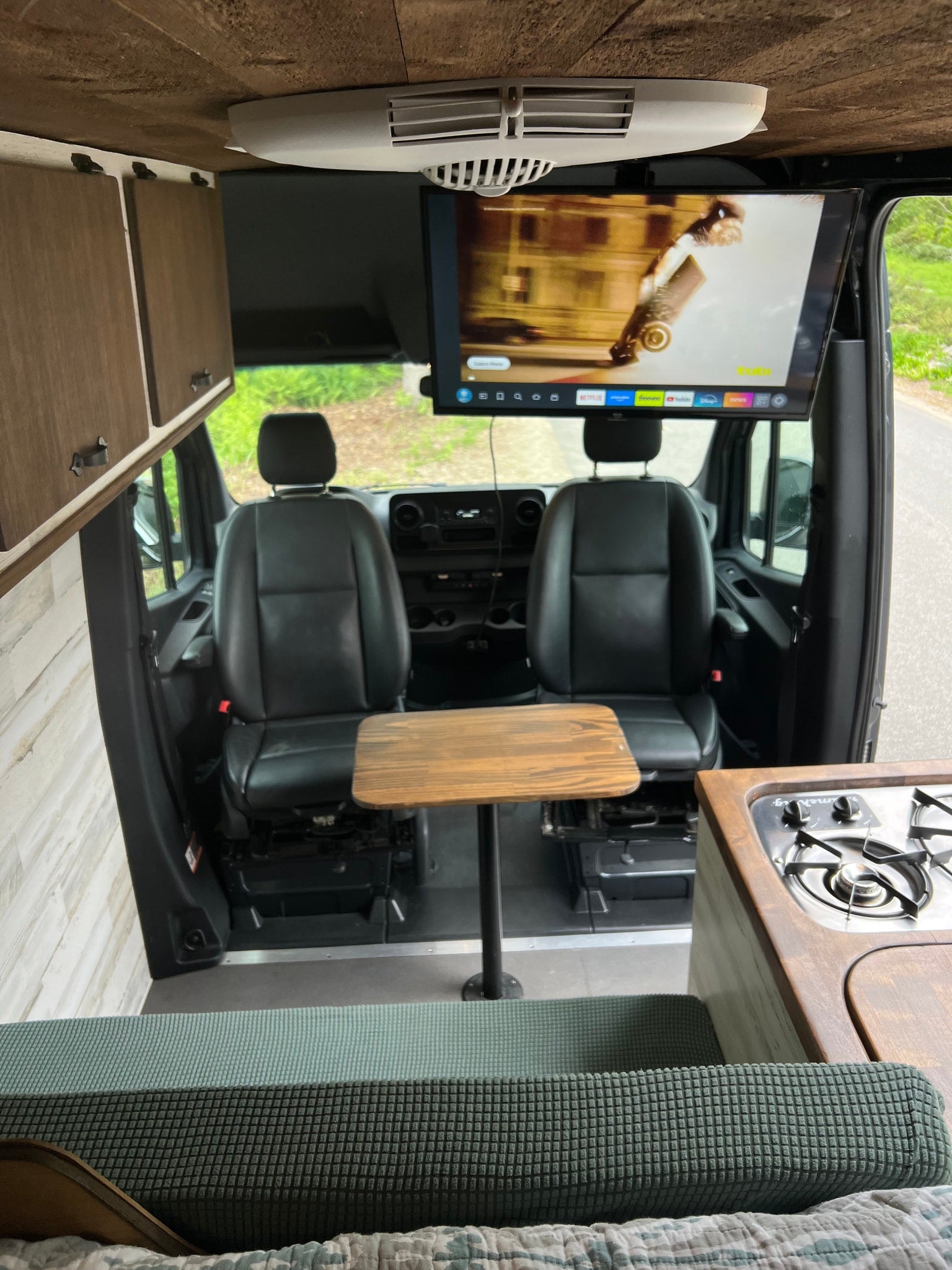 View inside an RV rental showing the front seats, a central table, an overhead TV, a kitchen counter with stove, and wooden cabinets.