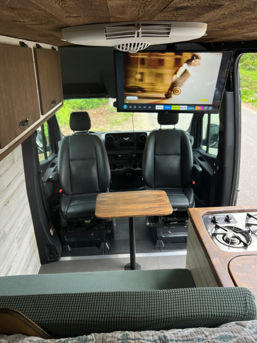 View inside an RV rental showing the front seats, a central table, an overhead TV, a kitchen counter with stove, and wooden cabinets.
