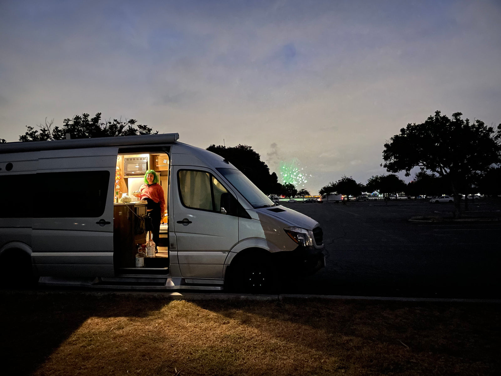 An RV rental provides a cozy space: A person cooks inside an illuminated camper van at night, with green fireworks in the distance.