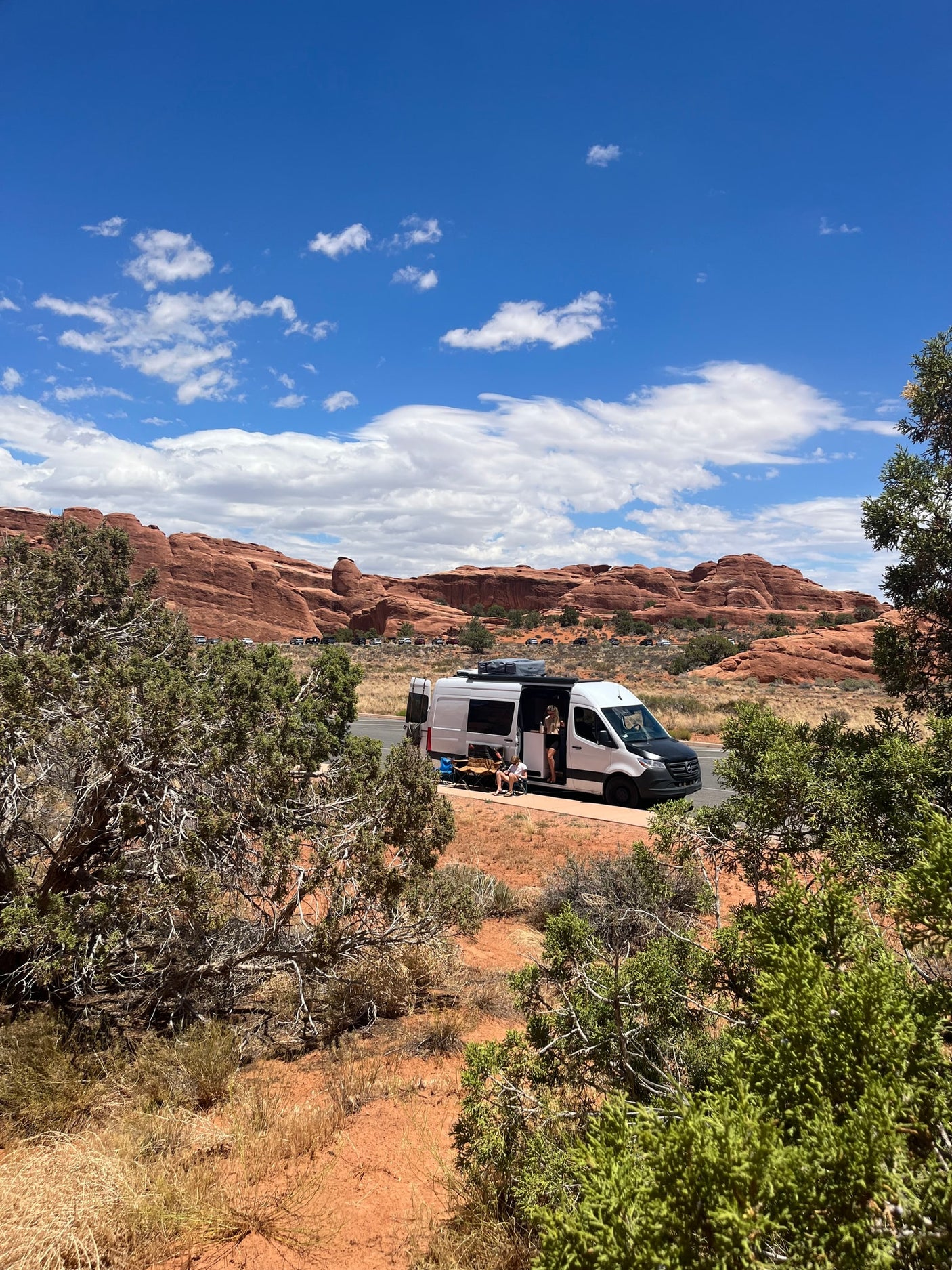 RV rental parked on desert road by red rock cliffs. Two people relax outside white camper van under blue, cloudy sky.