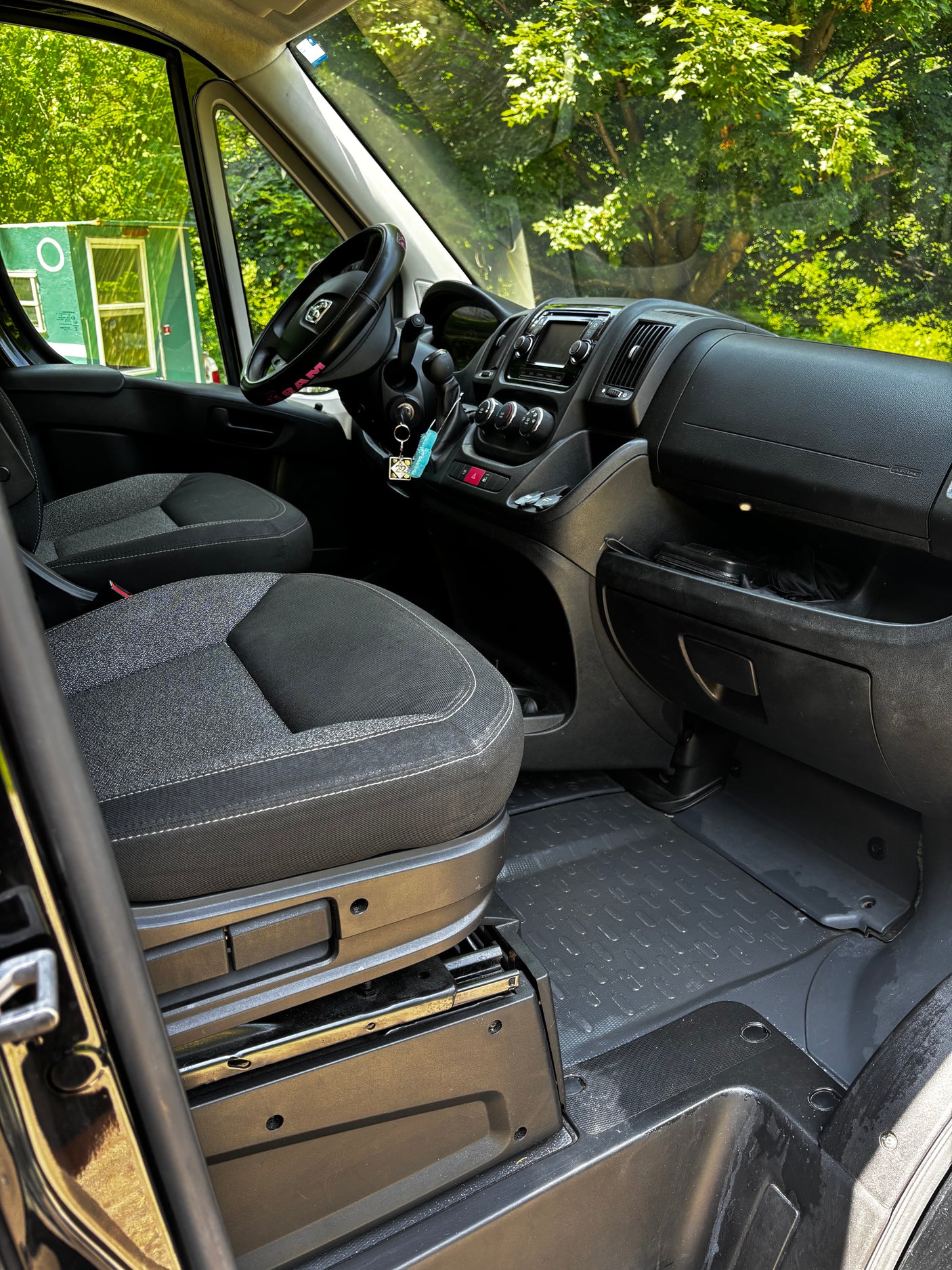 Interior of an RV rental van: clean, functional cabin with black and grey seats, dashboard, and a clear view of green trees.