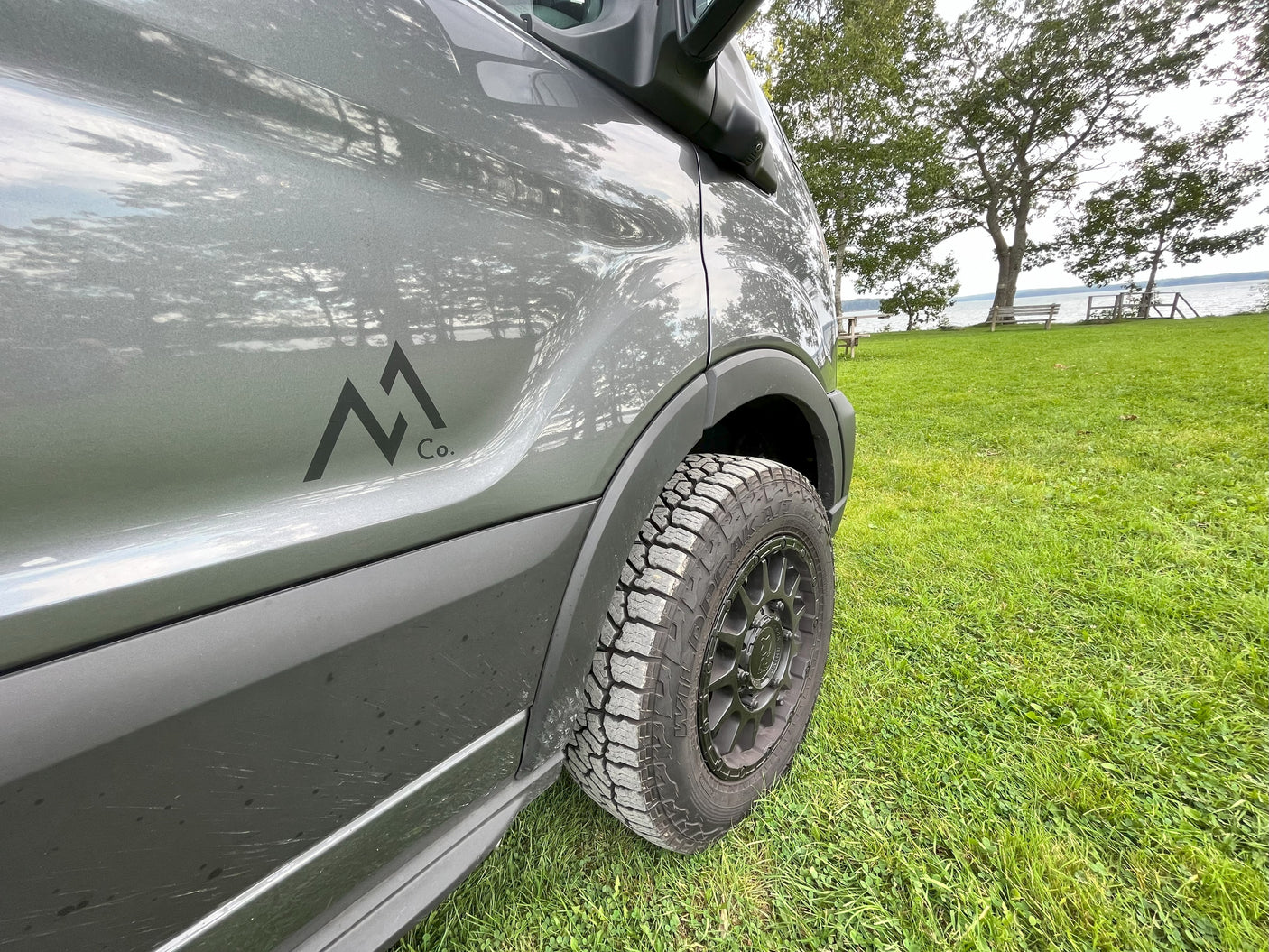 Adventure-ready gray RV rental van with an M1 Co. logo and rugged tires, parked on grass by a serene lake.