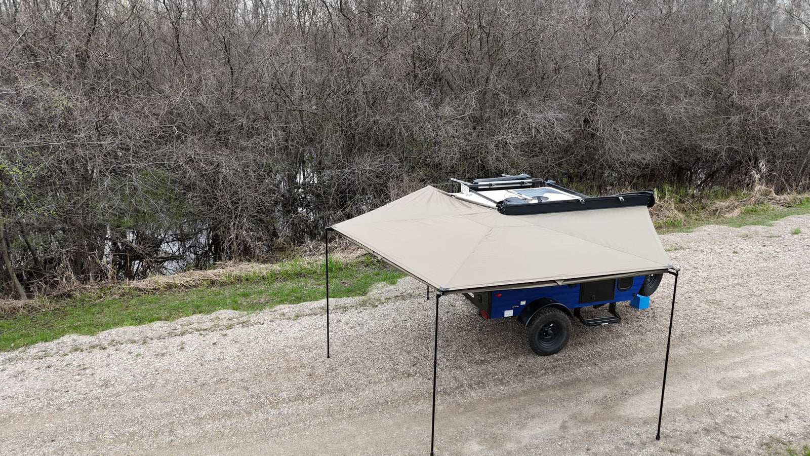 Blue off-road camper with extended awning on a gravel path, by dense, bare trees and water. Great RV rental for adventure.