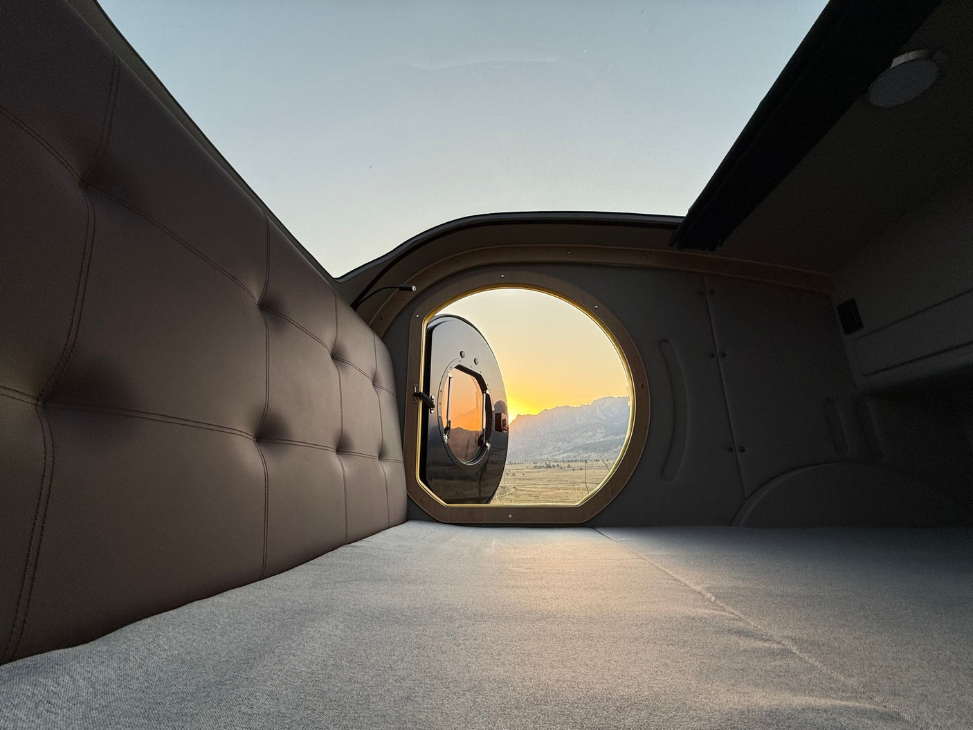 Comfortable rv rental interior: Tufted bed, unique round window open to a vibrant mountain sunset. Perfect for adventure.