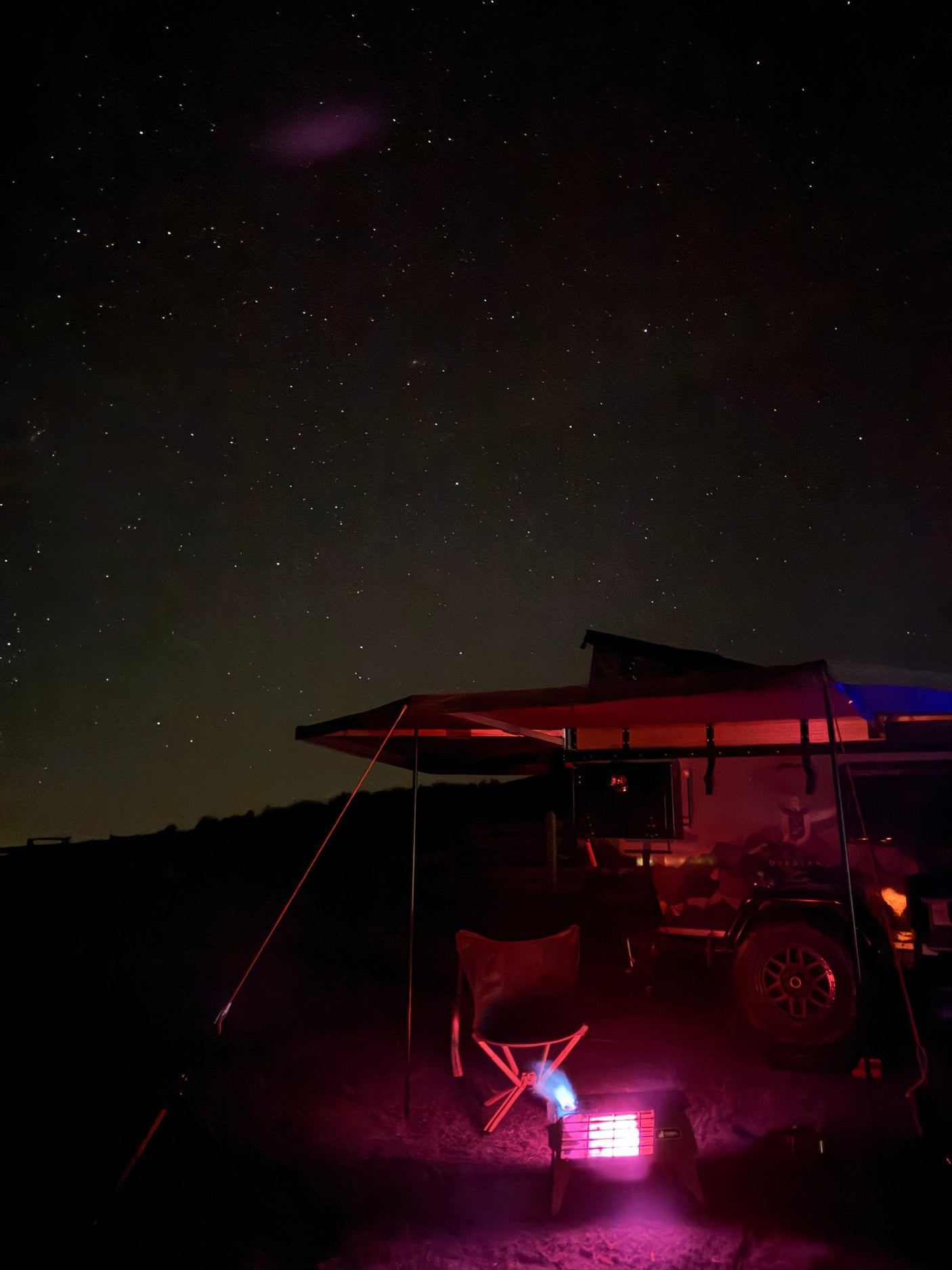 Experience starry nights! An rv rental with awning, chair, and glowing heater illuminates a cozy camp under a beautiful night sky.