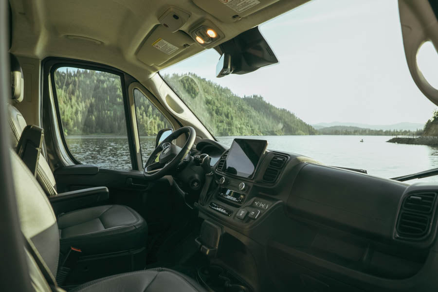RV rental camper van interior. Driver's seat & dashboard with large screen, overlooking a serene lake & forested mountains.
