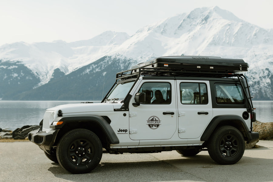 White Jeep Wrangler RV rental with rooftop tent by an Alaskan lake. Explore snowy mountains & wild landscapes.