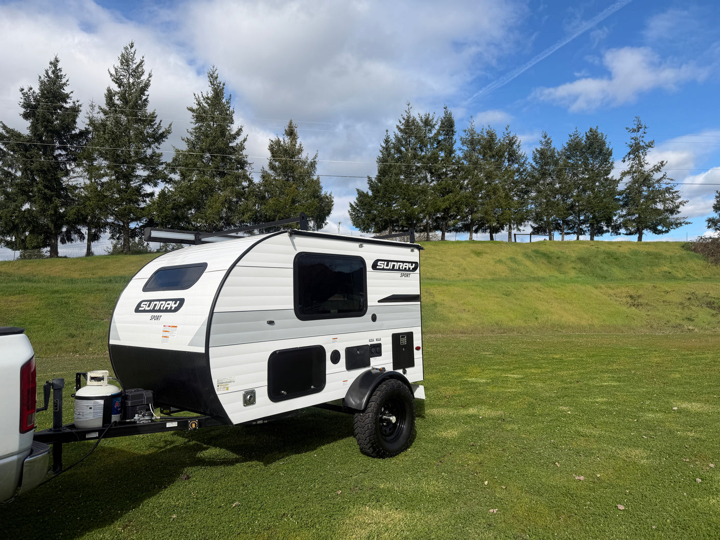 This Sunray Sport teardrop rv rental, hitched to a truck, awaits adventure on a lush green field under a blue sky.