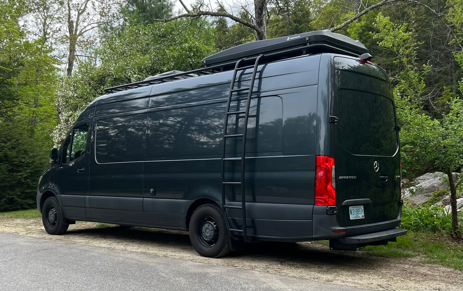 Side view of a modern, dark gray Mercedes Sprinter rv rental van with roof rack, rooftop tent, and side ladder.