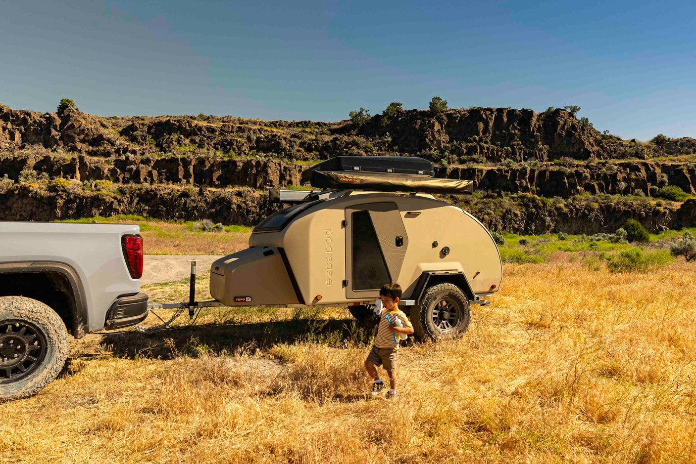 Adventure ready: A truck tows an Escapod teardrop RV rental on golden grass. Rocky bluffs and a boy walking complete the scene.