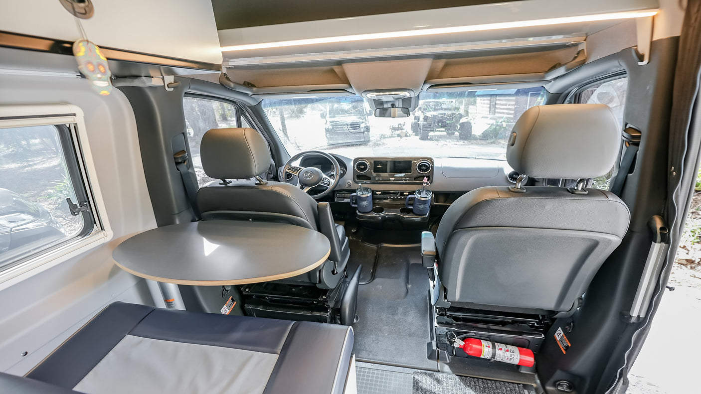 Inside an RV rental: a spacious camper van front cabin with swiveling seats, dashboard, and a dinette table. Fire extinguisher visible.