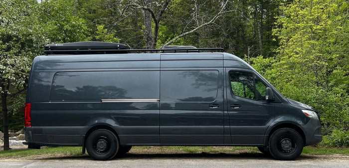 Side view of a sleek, dark grey camper van with roof cargo box & rack, set against green trees. Great for RV rental travel.