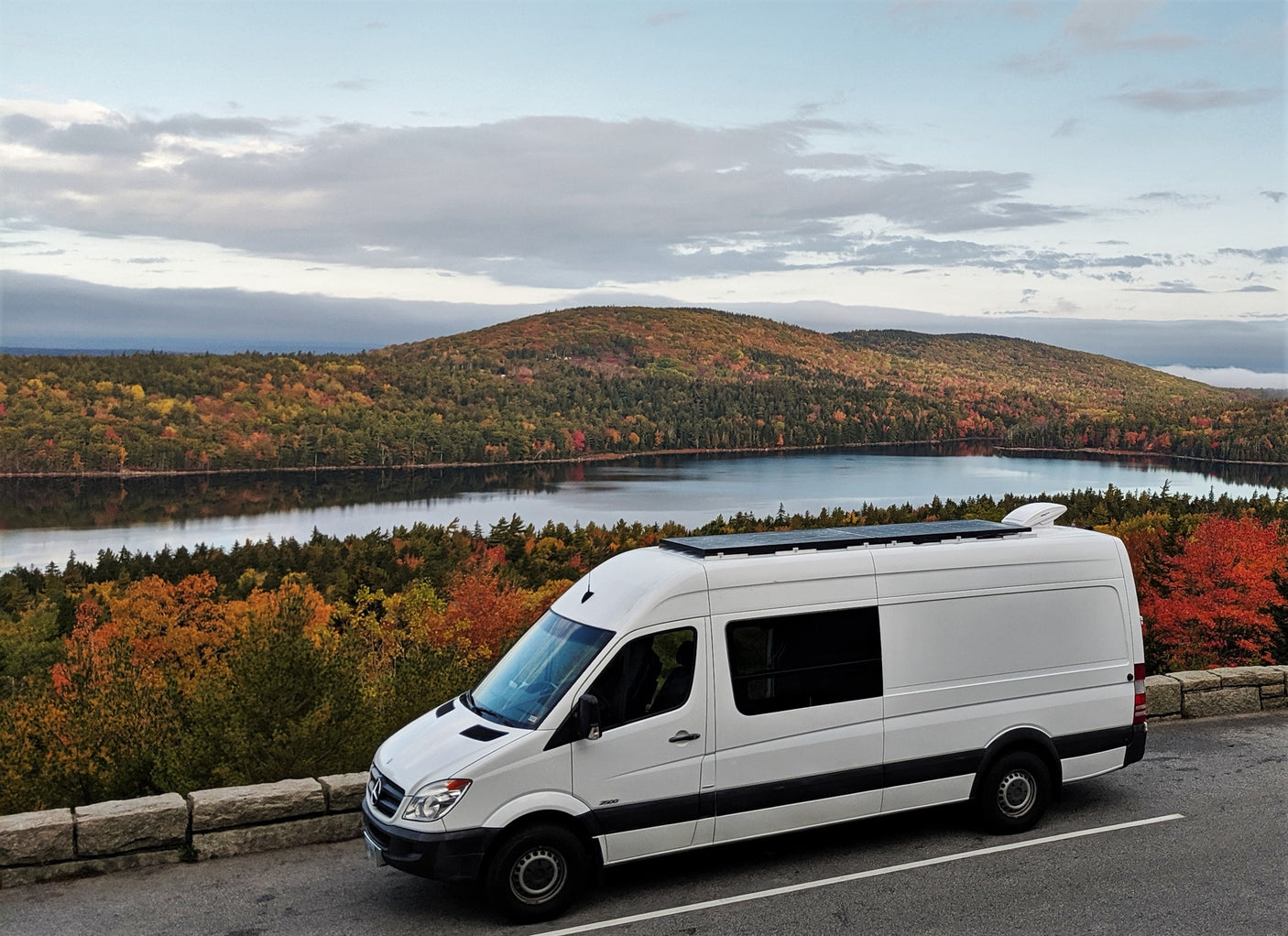 White rv rental camper van with solar panels, overlooking a stunning autumn lake and vibrant fall foliage.