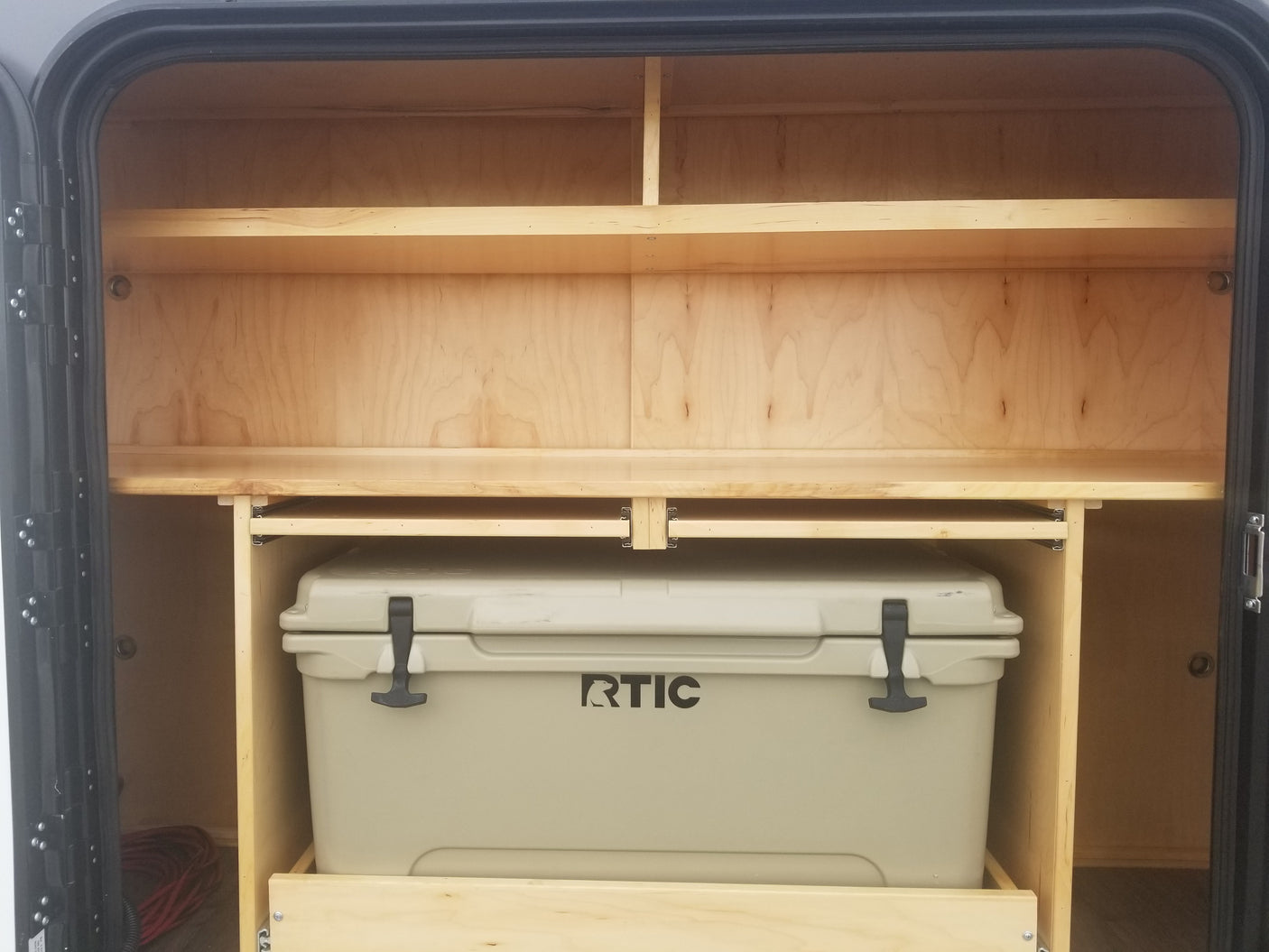 RV rental interior: organized wooden shelves above a pull-out tray holding an RTIC cooler, ideal for camping storage.