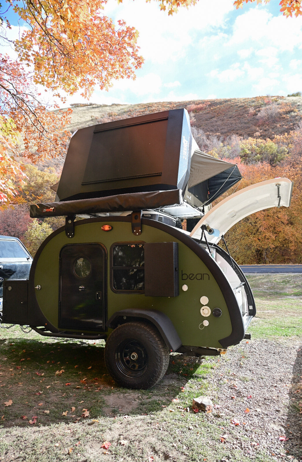 Green Bean teardrop trailer rv rental with rooftop tent, rear hatch open, parked in vibrant fall foliage. Ready for camping!