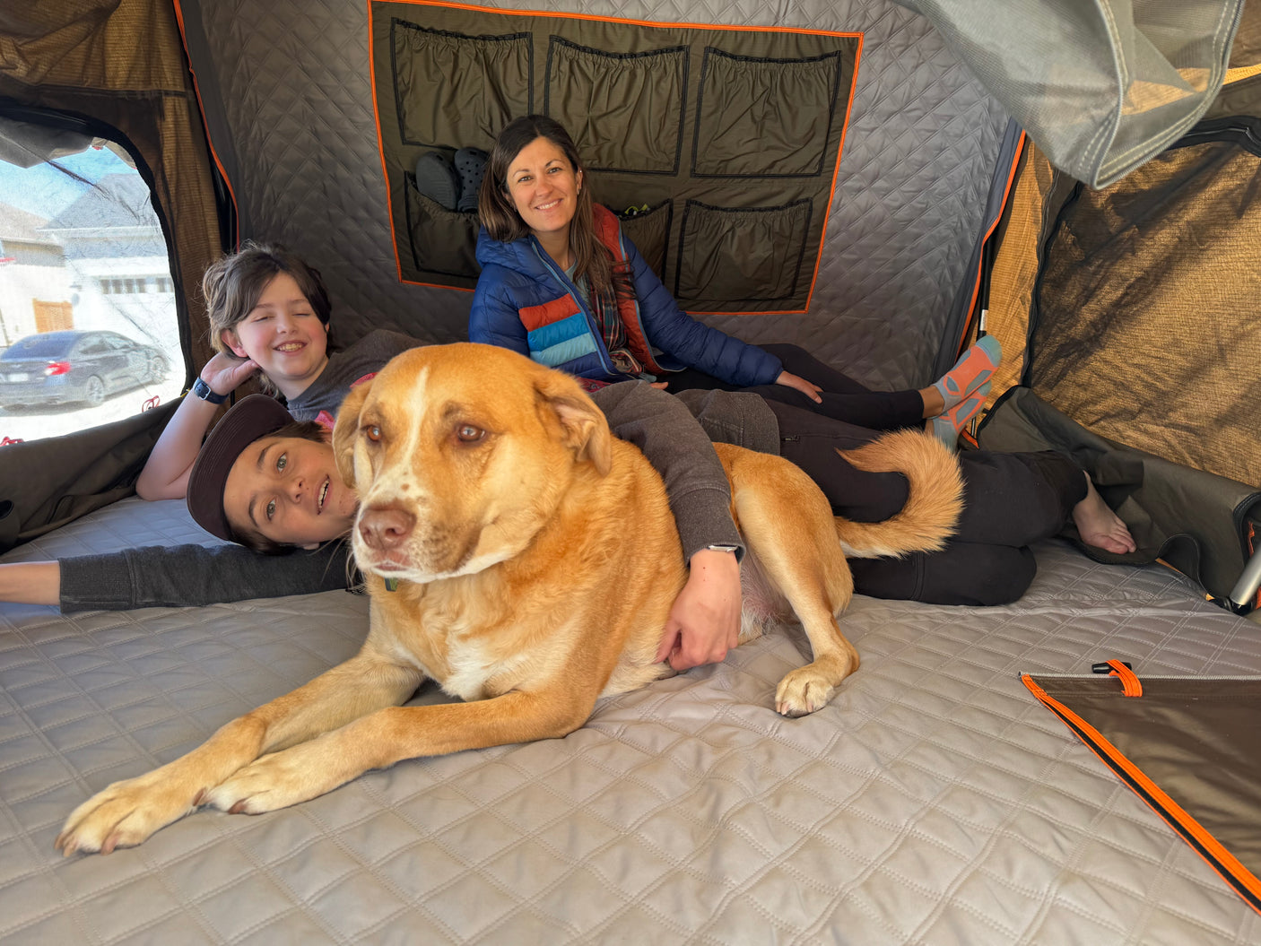 Cozy family fun in an RV rental's rooftop tent. Dog, two kids, and woman relax inside on a quilted mattress.