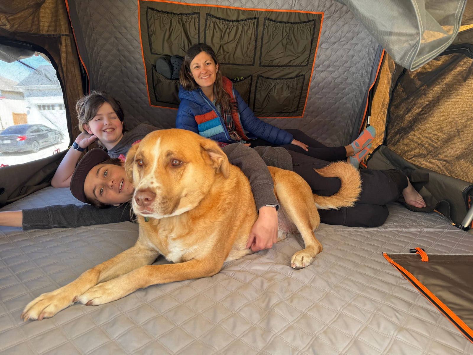 Cozy family fun in an RV rental's rooftop tent. Dog, two kids, and woman relax inside on a quilted mattress.
