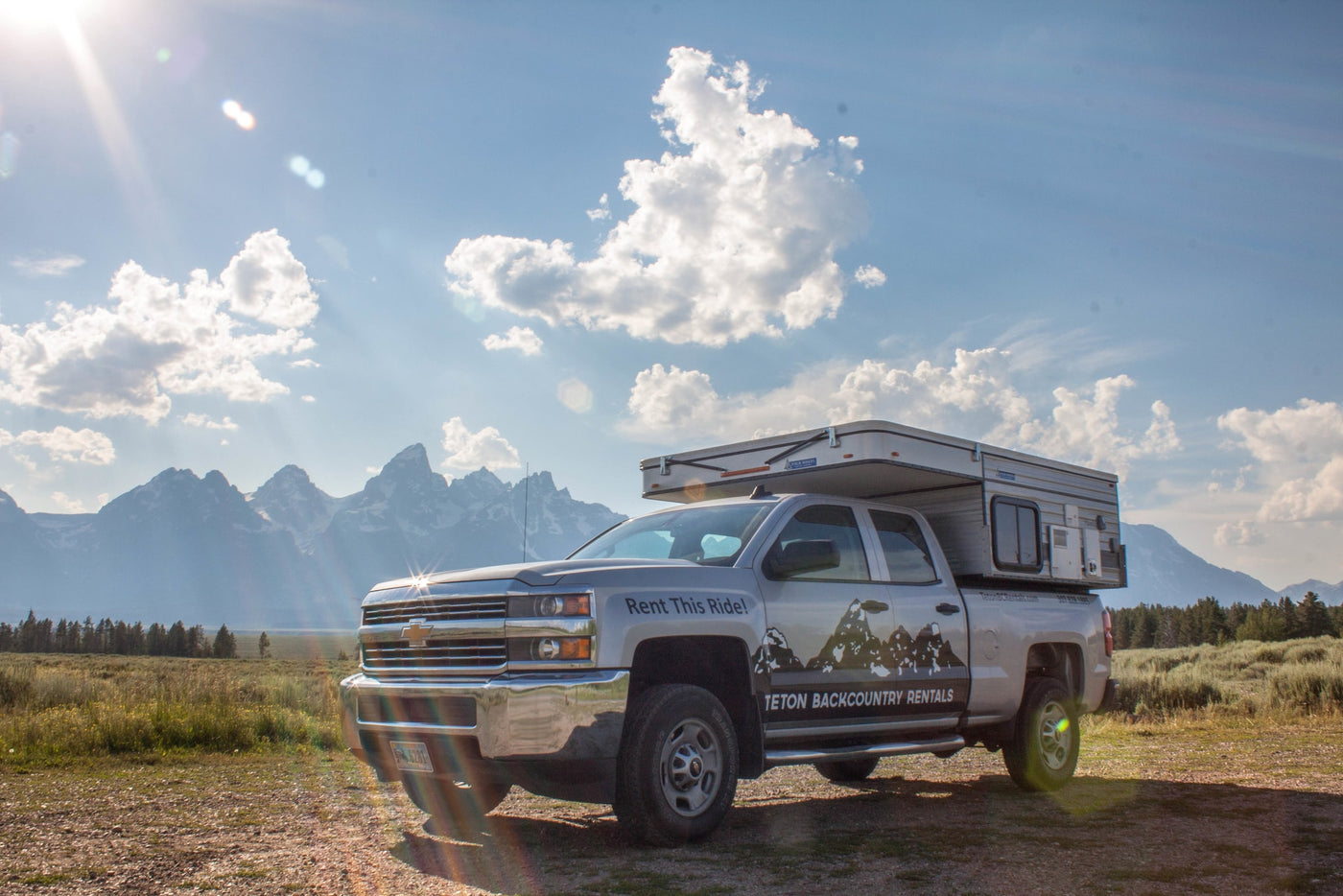 Explore the outdoors with this RV rental! A truck camper sits before the majestic Teton mountains under a bright sky. — photo 3