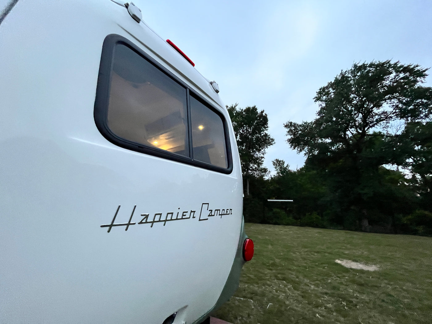 View of a white Happier Camper RV rental's rear, parked on grass at dusk, with interior lights glowing. — photo 5