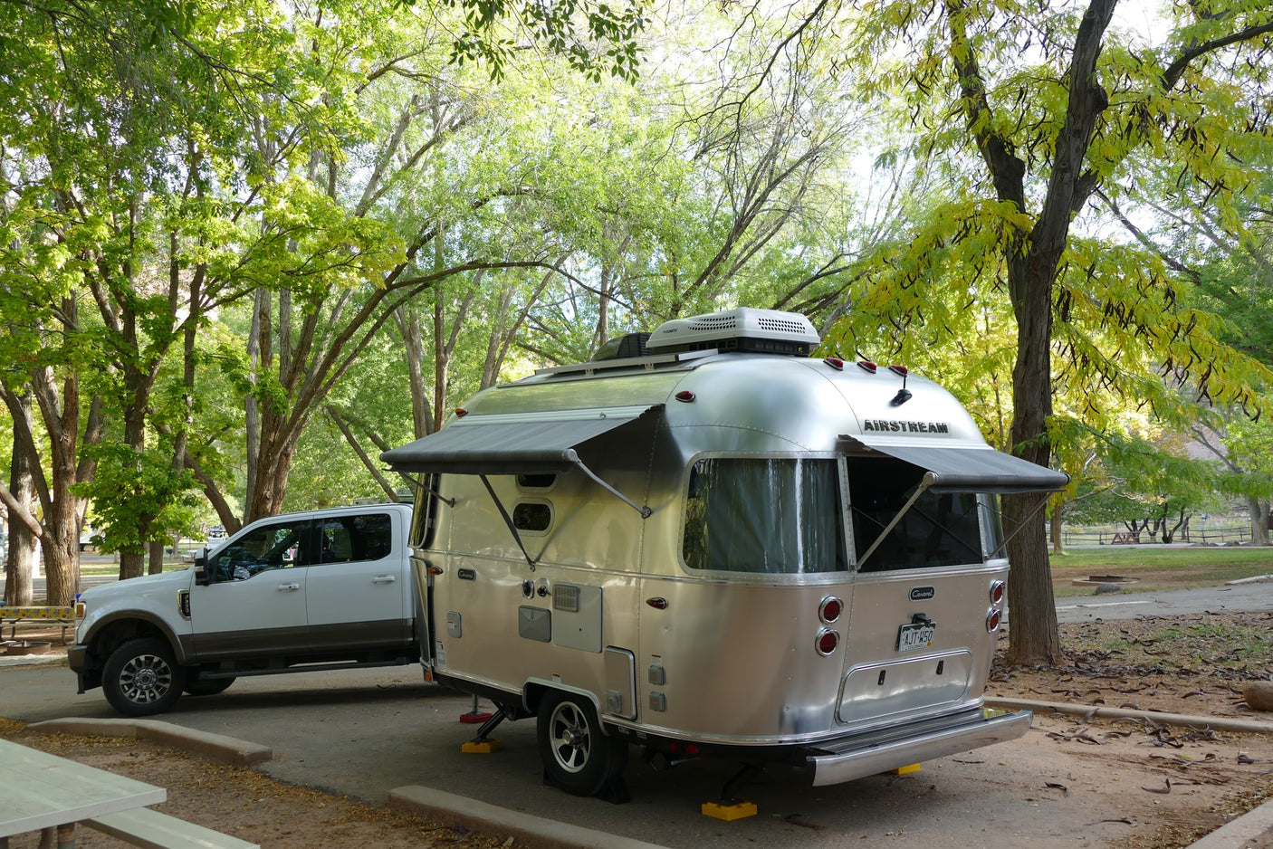 An Airstream RV rental, with awnings extended, is hitched to a white truck at a scenic, tree-filled campsite.