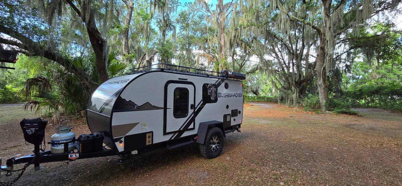 A Bushwhacker RV rental (rugged travel trailer) parked beneath majestic trees draped in Spanish moss at a serene campsite.