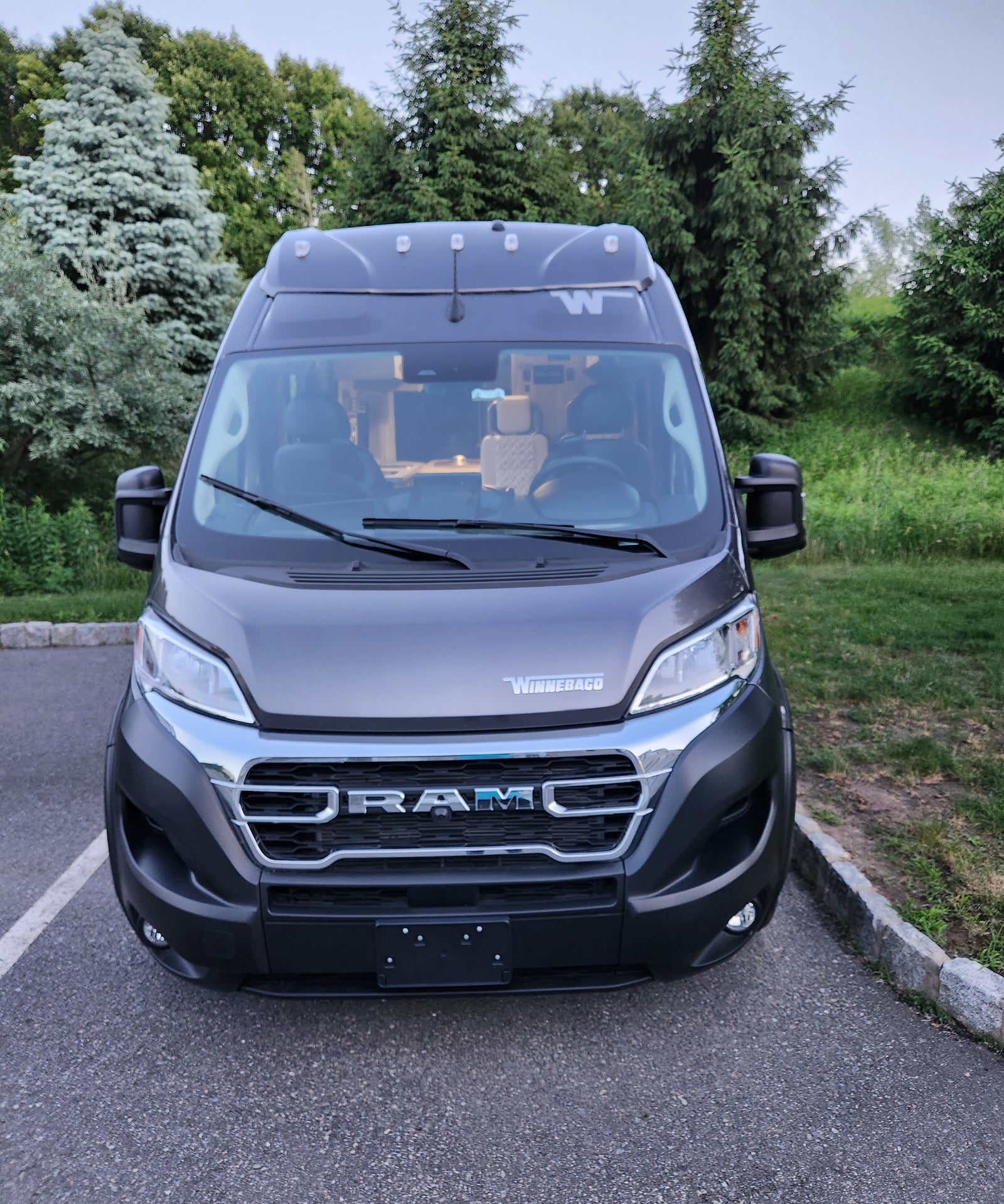 Front of a dark gray Winnebago Ram camper van, ideal for your next rv rental. Interior visible through windshield; parked by trees.