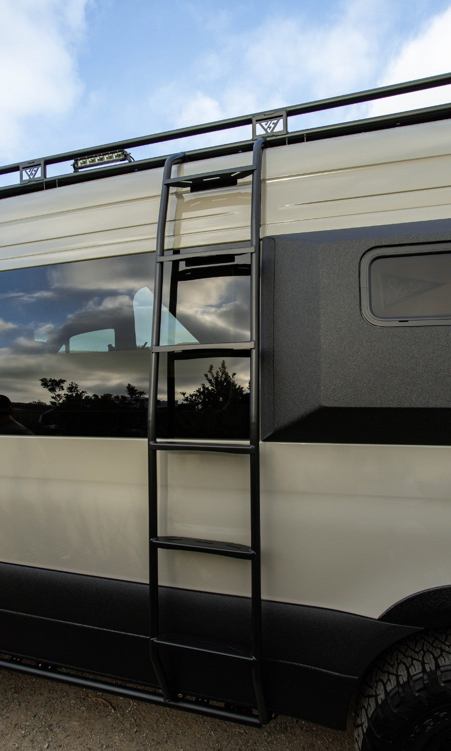 Close-up of a custom RV rental van, featuring a roof rack, sturdy access ladder, and rugged two-tone exterior.