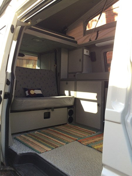 Cozy **RV rental** interior: bench seat/bed, cabinets, pop-top bunk, and colorful rugs from door view.