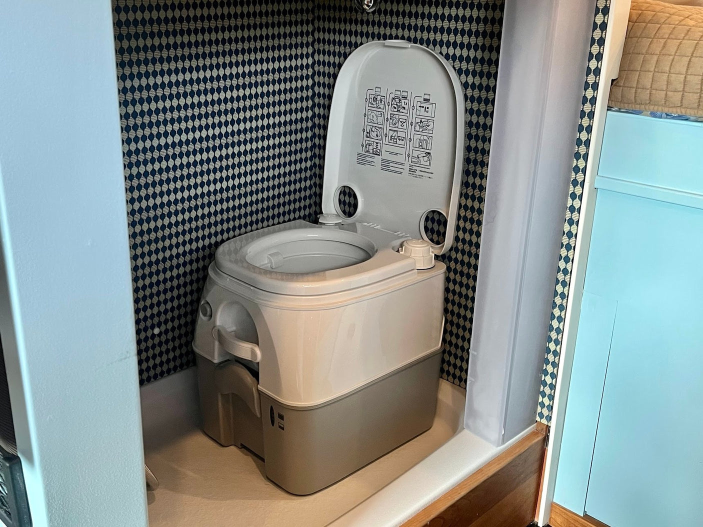 Portable toilet in an RV rental bathroom. The white and gray unit has its lid open, revealing operating instructions, set against patterned walls.