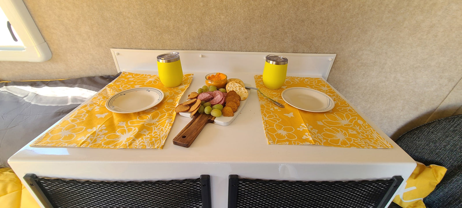 Charcuterie board in a cozy rv rental, with salami, grapes, crackers, dip, two plates & yellow cups on a white table.