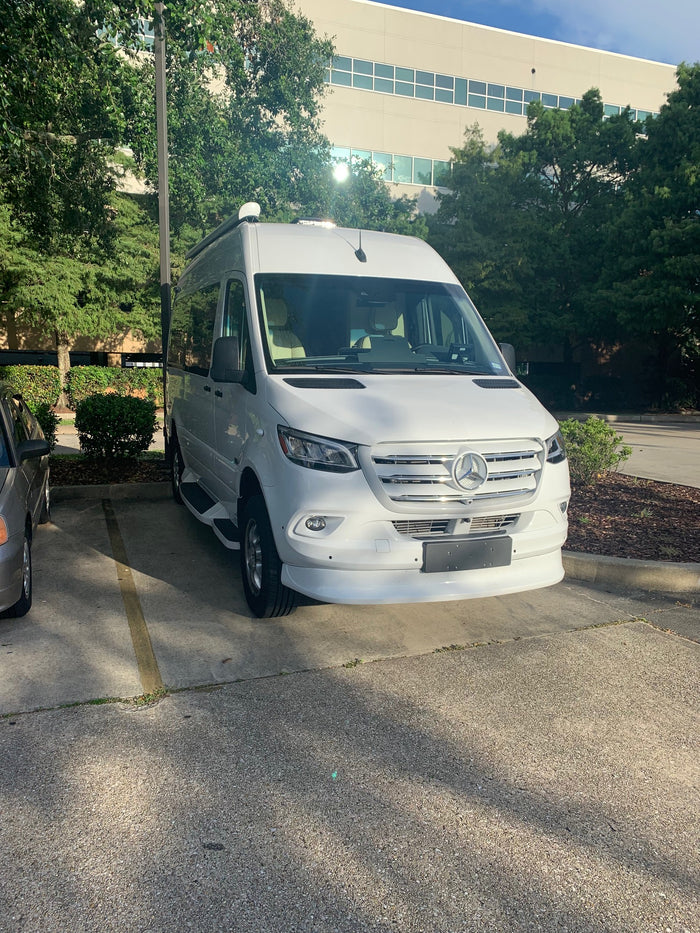 White Mercedes-Benz Sprinter van, an ideal RV rental, parked on concrete. Sunlight glints off its hood near a building and trees.